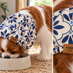 A brown and white cavalier dog wearing a blue and cream floral-patterned pet sood while eating from a white bowl. The image is a split-screen close-up showing the dog wearing the hood and a detailed view of the fabric pattern and a brown leather brand patch that reads 'TINKA pet'.