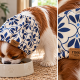 A brown and white cavalier dog wearing a blue and cream floral-patterned pet sood while eating from a white bowl. The image is a split-screen close-up showing the dog wearing the hood and a detailed view of the fabric pattern and a brown leather brand patch that reads 'TINKA pet'.
