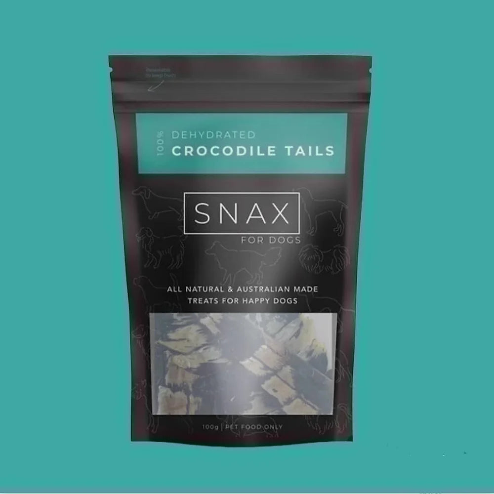 A package of SNAX brand dried crocodile tails for dogs, presented against a white background with a visible clear window showing the tails inside the package.