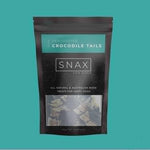 A package of SNAX brand dried crocodile tails for dogs, presented against a white background with a visible clear window showing the tails inside the package.