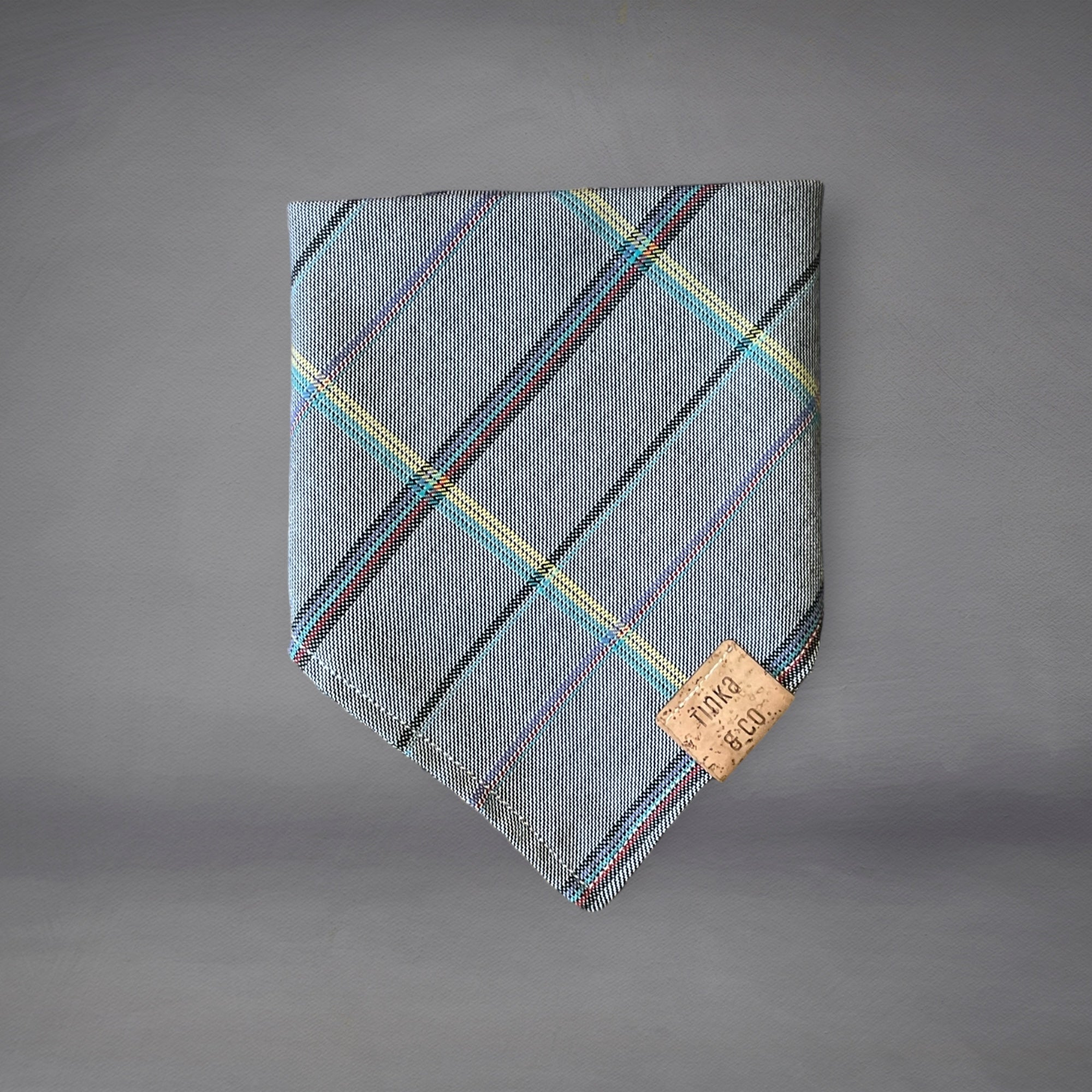 A gray plaid dog bandana with multicolor lines, displayed on a flat surface with a brand tag visible.
