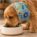 A Cocker Spaniel dog eating from a speckled beige ceramic bowl while wearing a colorful floral Snood.  The snood features a turquoise blue background with orange, purple, and red floral patterns, and has a brown leather patch with the brand name 'TINKA & CO'.