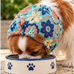 A Cavalier dog wearing a colorful floral snood while drinking from a white bowl with blue paw prints. The hat has a turquoise, orange, and navy blue floral pattern with a small brown leather patch that reads 'TINKA & CO'.