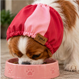 A small brown and white dog wearing a pink and red petSnood with a TINKA & CO label, eating from a pink dog bowl.