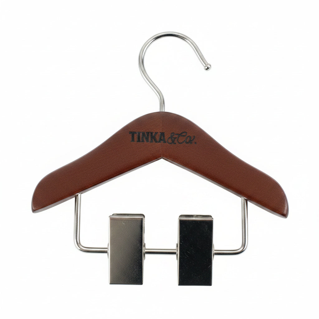 A wooden clothes hanger with a brown finish and silver metal clips and hook, featuring the brand name 'TINKA & Co.' printed on the front, set against a textured white background.