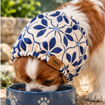 A brown and white dog wearing a blue and cream floral patterned pet snood while eating from a blue bowl outdoors. The hat has a small brown leather patch with the brand name 'TINKA & CO'